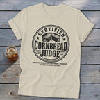 Cream t-shirt with a black graphic reading Certified Cornbread Judge and a cornbread loaf icon, laid on wooden boards.