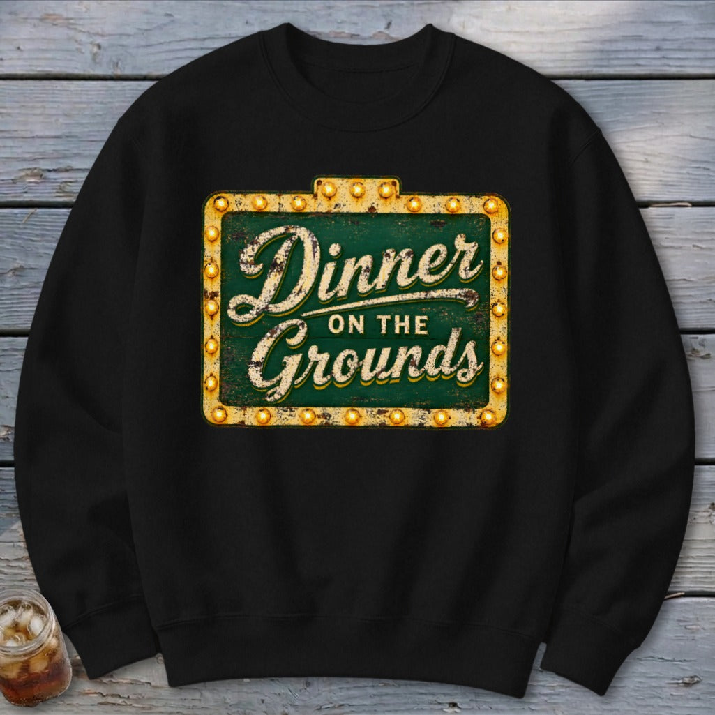 A black crewneck sweatshirt featuring a distressed green marquee sign graphic with the text Dinner on the Grounds.