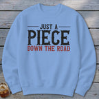 Light blue sweatshirt with distressed text "JUST A PIECE DOWN THE ROAD" in black and red.