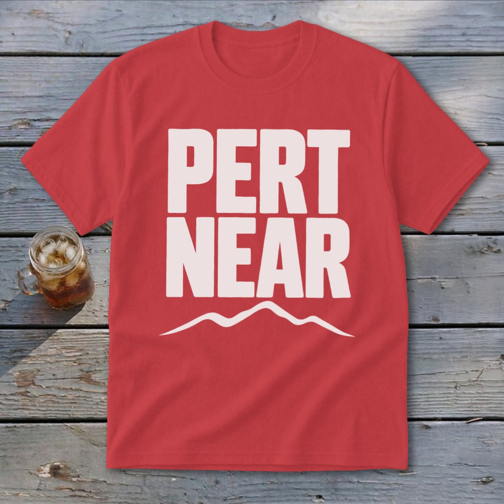 A red short-sleeved t-shirt with white block text reading PERT NEAR above a mountain outline, lying on wooden planks beside a glass of iced tea.