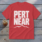 A red short-sleeved t-shirt with white block text reading PERT NEAR above a mountain outline, lying on wooden planks beside a glass of iced tea.