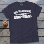 A navy blue t-shirt with white text reading "THIS THANKSGIVING IS SPONSORED BY SOUP BEANS" lies on a wooden background next to a glass of iced tea.