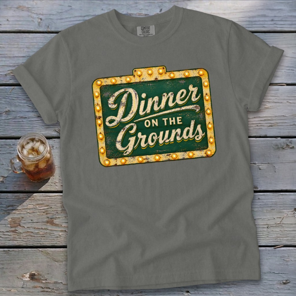 A grey t-shirt featuring a green vintage marquee sign graphic that reads Dinner on the Grounds.