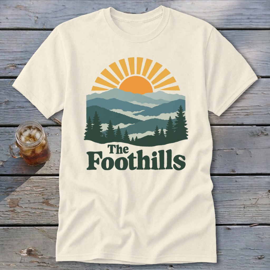 Cream t-shirt with sun setting over layered mountains and pine trees, reads "The Foothills".