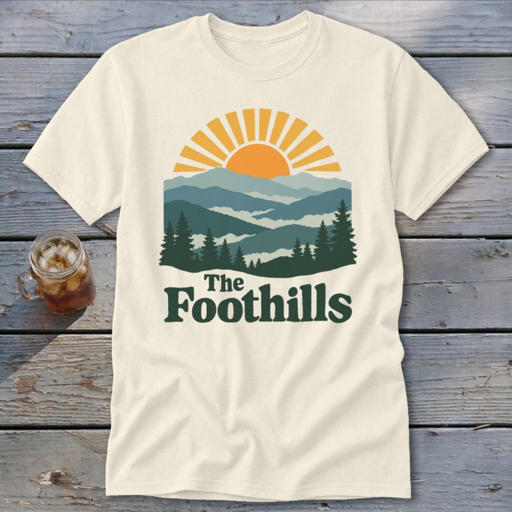 Cream t-shirt with sun setting over layered mountains and pine trees, reads "The Foothills".