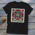A black short-sleeved t-shirt featuring a red, black, and white geometric quilt block graphic with the text "APPALACHIA" below it, lying on a wooden surface beside a glass of iced tea.
