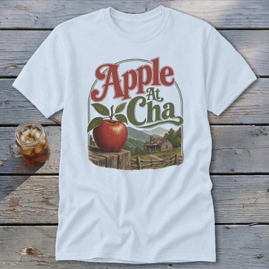 A white t-shirt featuring a retro graphic of an apple on a fence before a cabin and mountains with the text "Apple At Cha".