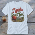 A white t-shirt featuring a retro graphic of an apple on a fence before a cabin and mountains with the text "Apple At Cha".