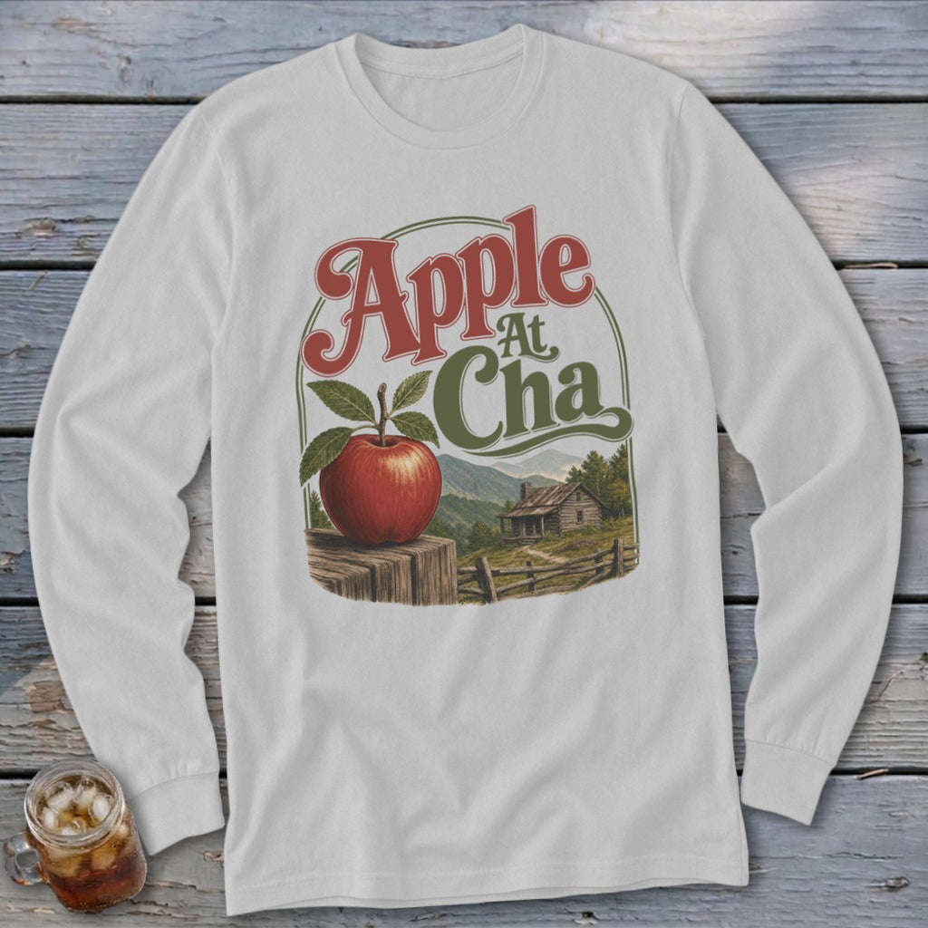 A light gray long-sleeve t-shirt featuring a graphic of a red apple, a cabin, and text reading Apple At Cha.