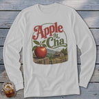 A light gray long-sleeve t-shirt featuring a graphic of a red apple, a cabin, and text reading Apple At Cha.