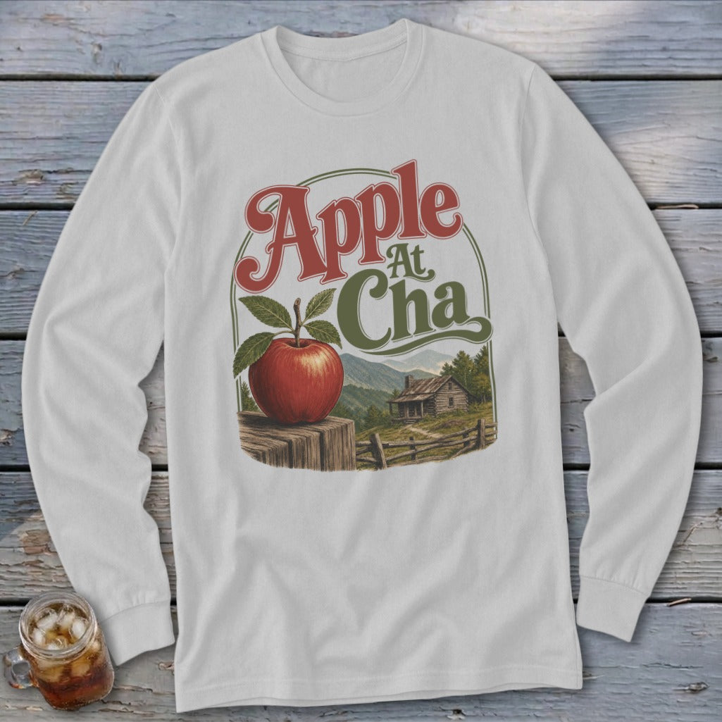A light gray long-sleeve t-shirt featuring a graphic of a red apple, a cabin, and text reading Apple At Cha.