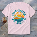 Light pink t-shirt with a retro round graphic depicting mountains, a sun, and text saying Born in the Holler Raised on the Ridge.