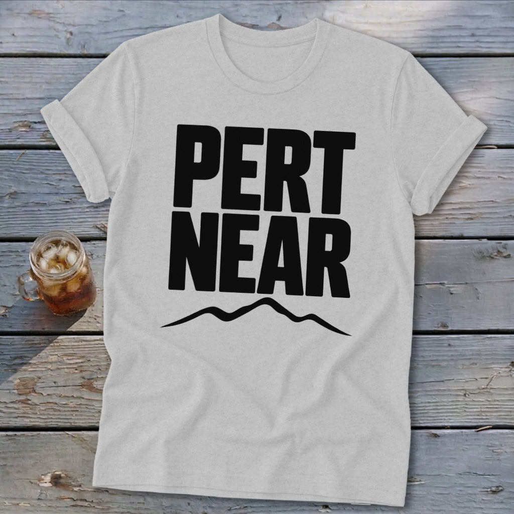 Light gray crewneck t-shirt featuring the black text PERT NEAR above a mountain range graphic, resting on a weathered wooden surface with a glass of iced tea.