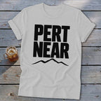 Light gray crewneck t-shirt featuring the black text PERT NEAR above a mountain range graphic, resting on a weathered wooden surface with a glass of iced tea.
