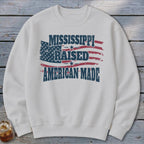 A cream-colored crewneck sweatshirt featuring a distressed American flag graphic and text reading MISSISSIPPI RAISED AMERICAN MADE.