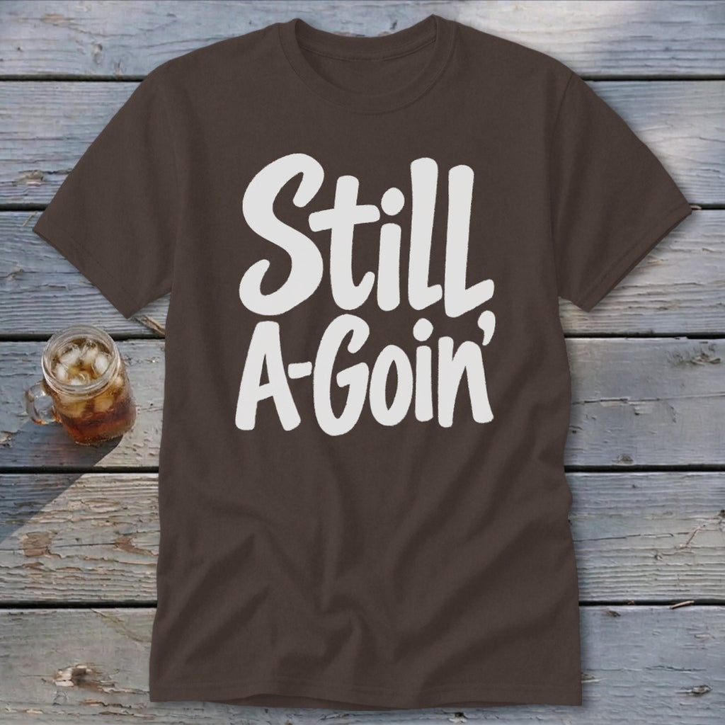 Brown t-shirt featuring the white text "Still A-Goin'" laid flat on a rustic wooden surface with a glass of iced tea.