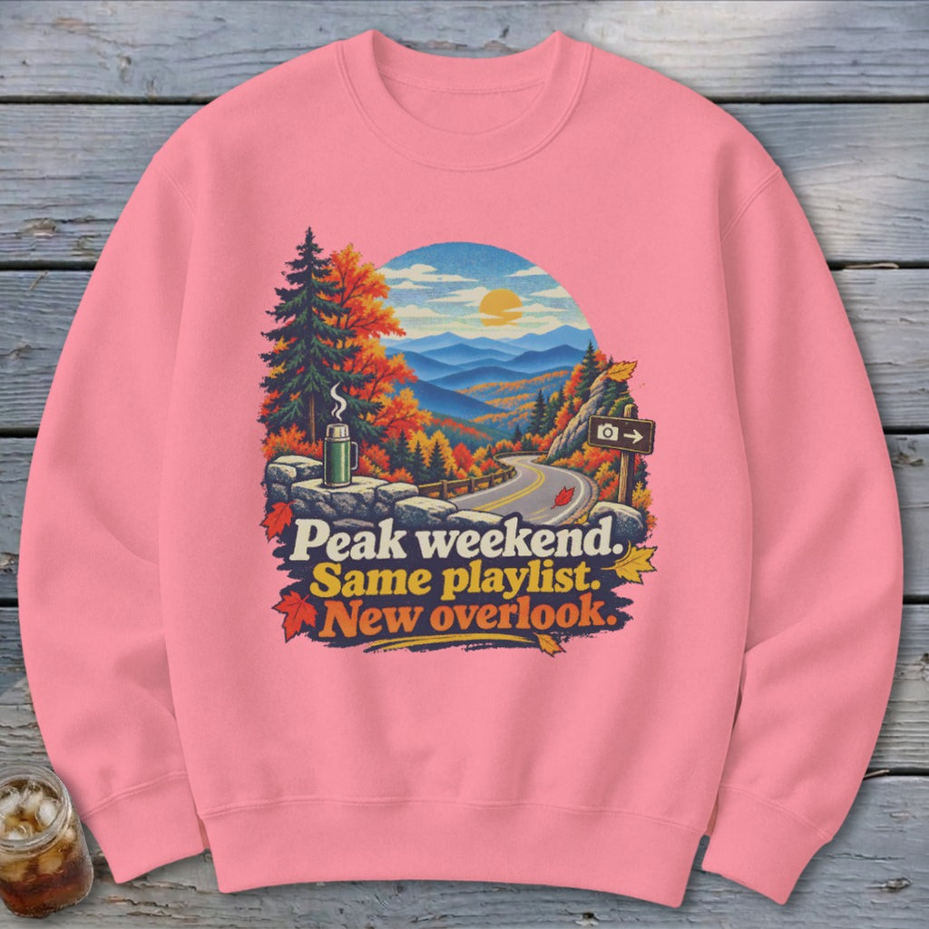 Pink sweatshirt with a graphic of autumn mountains, a road, and text: Peak weekend. Same playlist. New overlook.