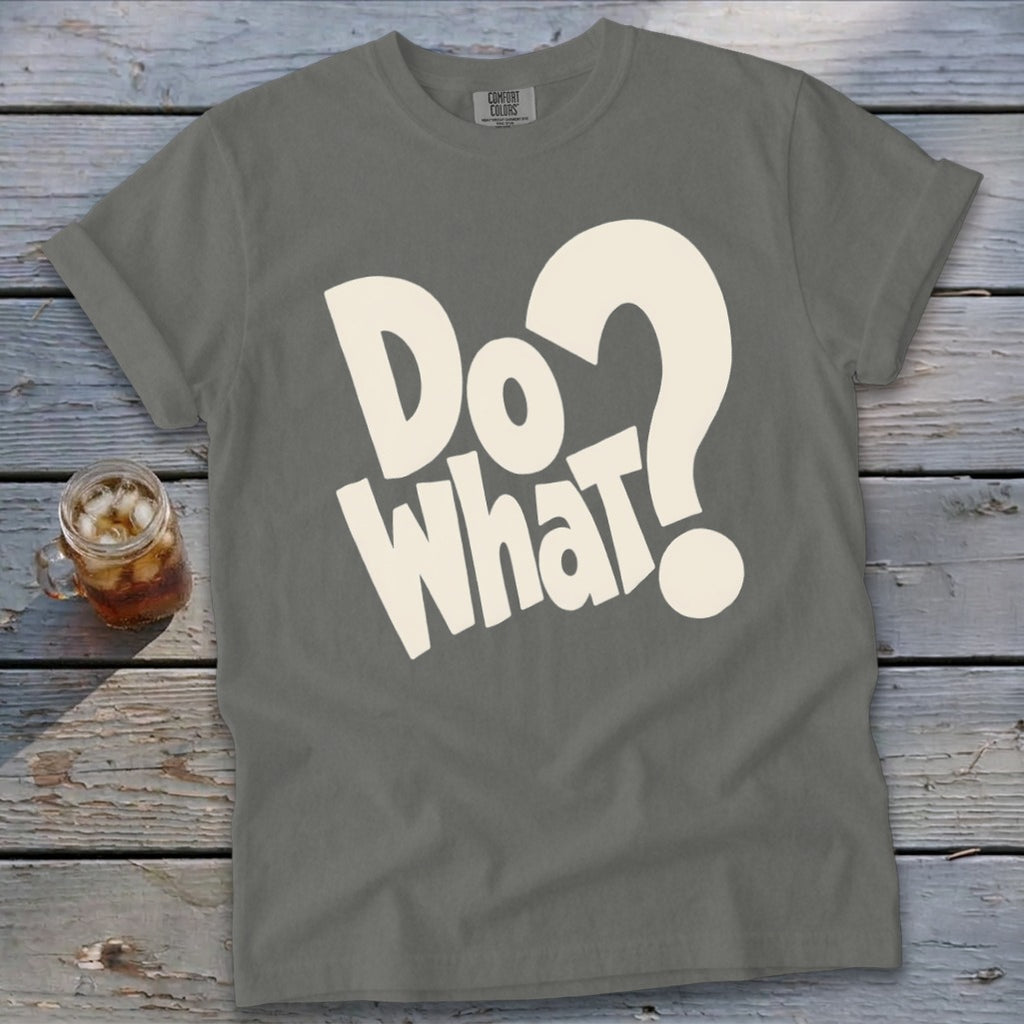 A gray crewneck t-shirt with large white lettering that reads Do What? laid flat on a weathered wooden surface next to a glass of iced tea.