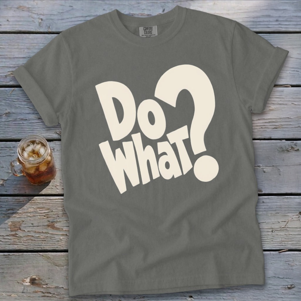 A gray crewneck t-shirt with large white lettering that reads Do What? laid flat on a weathered wooden surface next to a glass of iced tea.