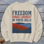 A beige crewneck sweatshirt features the text Freedom Rings Loudest In These Hills above a stylized mountain graphic.
