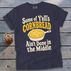 A dark blue t-shirt with graphic text reading "Some of Y’all’s Cornbread Ain’t Done in the Middle" and a frying pan with cornbread.