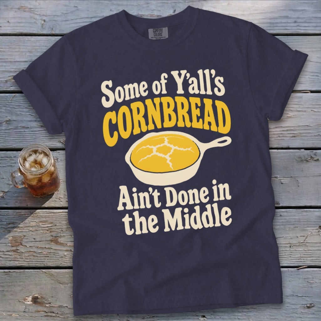 A dark blue t-shirt with graphic text reading "Some of Y’all’s Cornbread Ain’t Done in the Middle" and a frying pan with cornbread.