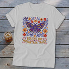 White t-shirt with a purple moth, flowers, bird, and text "BELIEVE THE MOUNTAIN TALES".