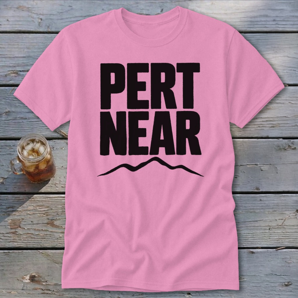 A pink short-sleeve t-shirt with the black text "PERT NEAR" and a mountain silhouette underneath, laid on a wooden background.