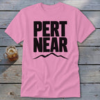 A pink short-sleeve t-shirt with the black text "PERT NEAR" and a mountain silhouette underneath, laid on a wooden background.