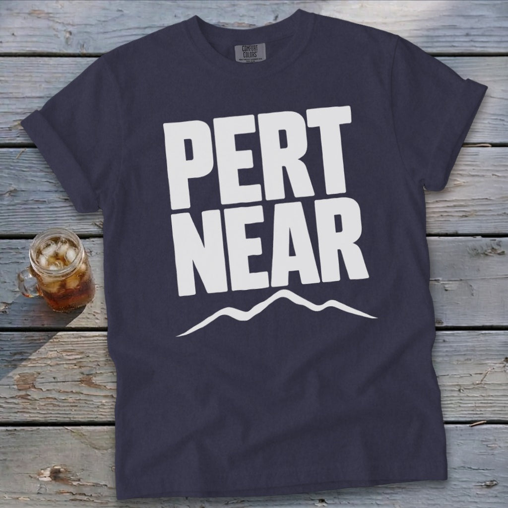 Dark blue t-shirt featuring the white text PERT NEAR above a mountain range graphic on a wooden surface.