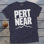 Dark blue t-shirt featuring the white text PERT NEAR above a mountain range graphic on a wooden surface.