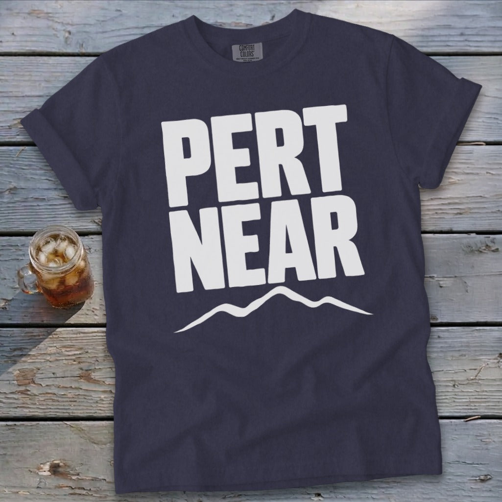 Dark blue t-shirt featuring the white text PERT NEAR above a mountain range graphic on a wooden surface.