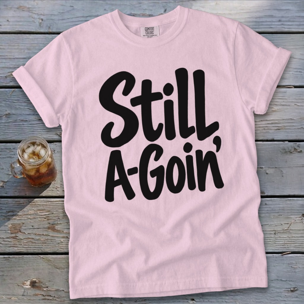Light pink t-shirt with black text that reads Still A-Goin, displayed on a wooden surface.