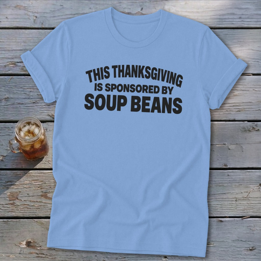 A light blue t-shirt printed with the black text THIS THANKSGIVING IS SPONSORED BY SOUP BEANS rests on wooden boards alongside a glass of iced tea.