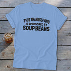 A light blue t-shirt printed with the black text THIS THANKSGIVING IS SPONSORED BY SOUP BEANS rests on wooden boards alongside a glass of iced tea.
