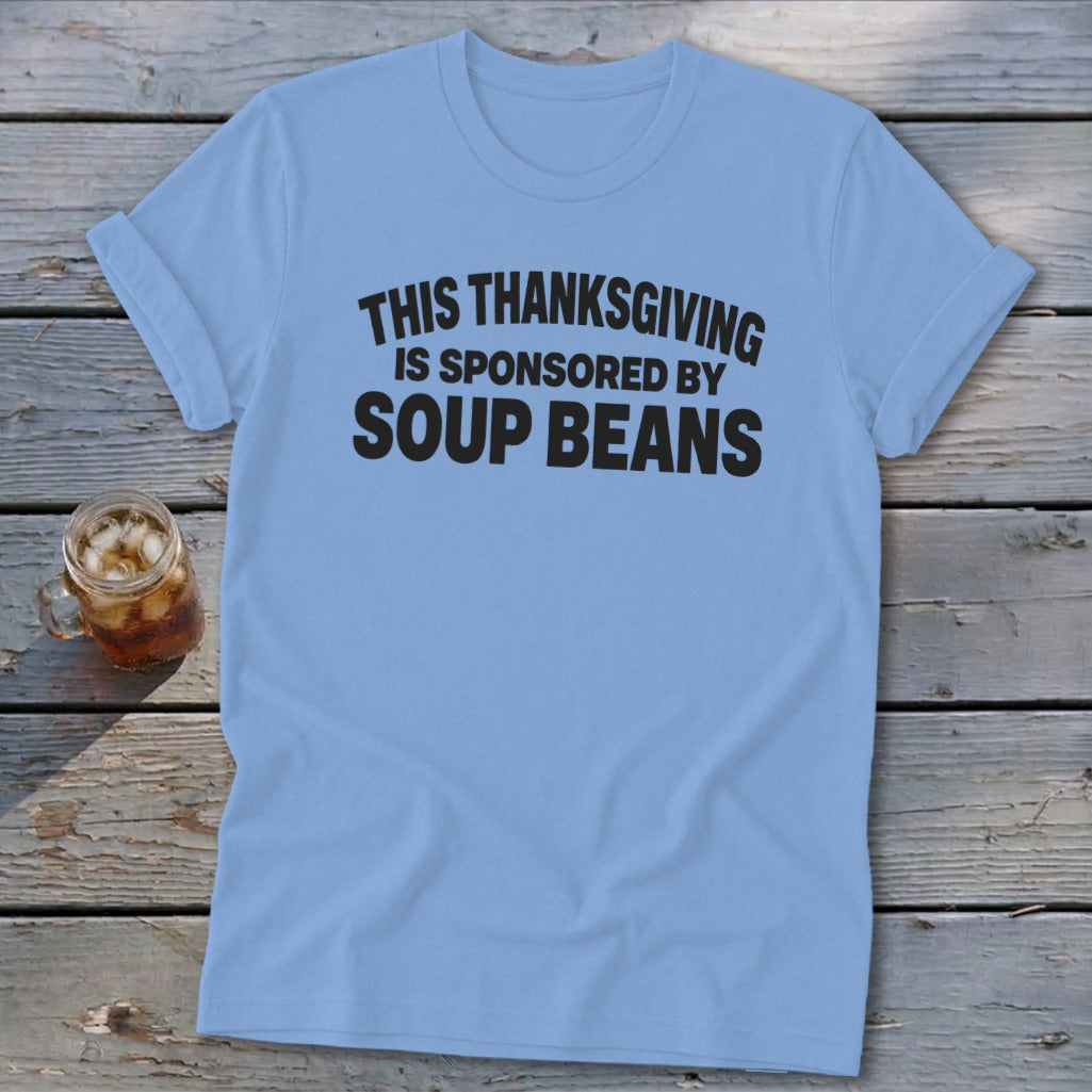 A light blue t-shirt printed with the black text THIS THANKSGIVING IS SPONSORED BY SOUP BEANS rests on wooden boards alongside a glass of iced tea.