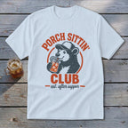 A white t-shirt featuring a retro graphic of a bear drinking iced tea with the text Porch Sittin Club.