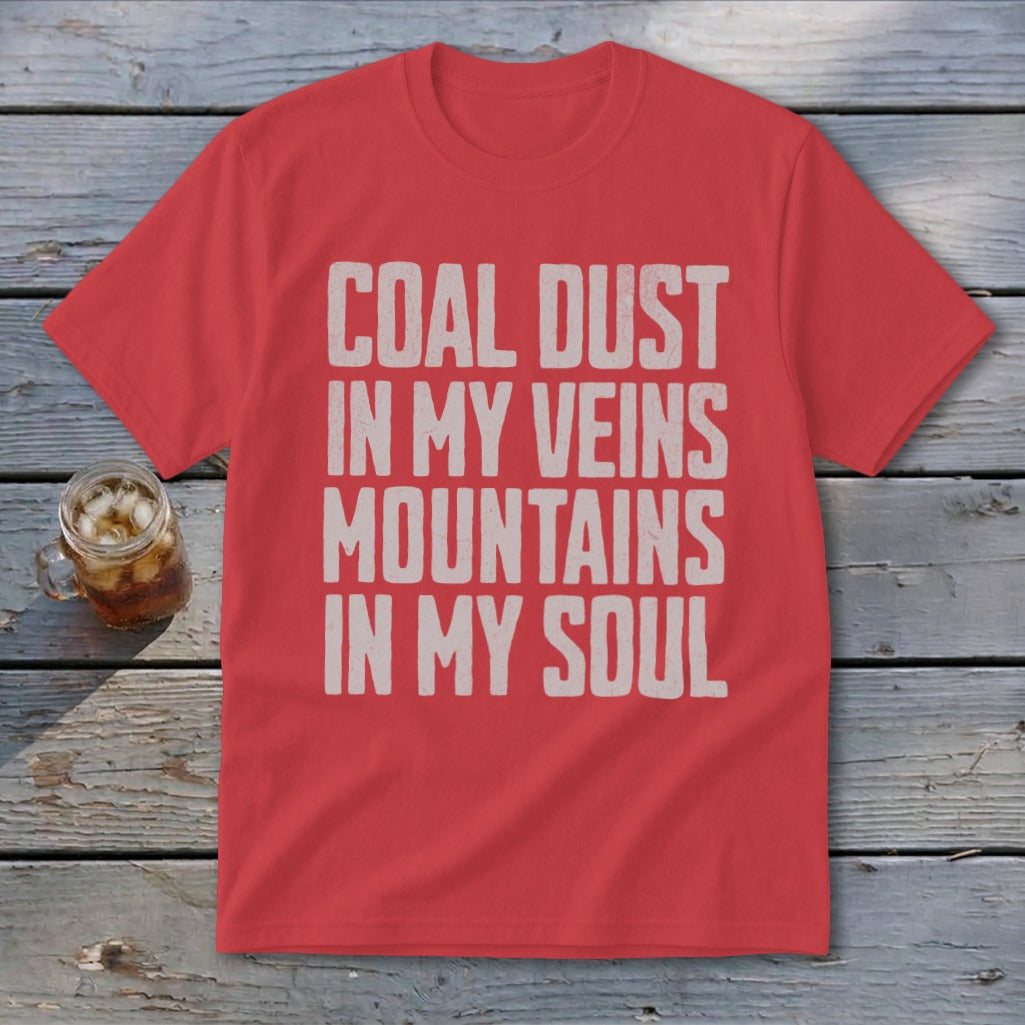 A red crewneck t-shirt featuring distressed white text that reads COAL DUST IN MY VEINS MOUNTAINS IN MY SOUL, laid on weathered wood next to a glass of iced tea.