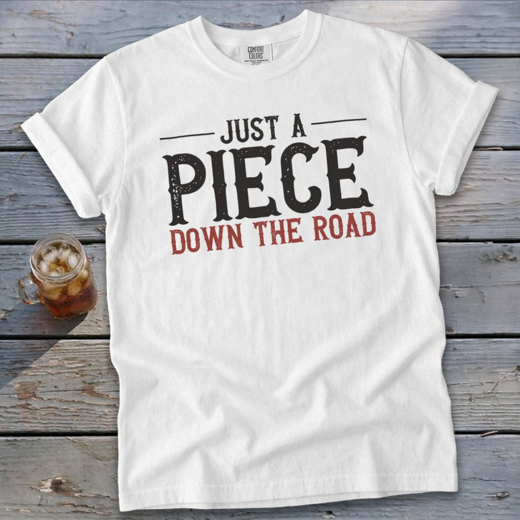 White t-shirt with text "JUST A PIECE DOWN THE ROAD" and a drink.
