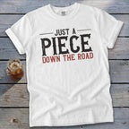 White t-shirt with text "JUST A PIECE DOWN THE ROAD" and a drink.