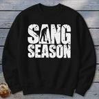 A black long-sleeved crewneck sweatshirt with white distressed text that reads SANG SEASON, featuring a person digging with a shovel inside the letter A.