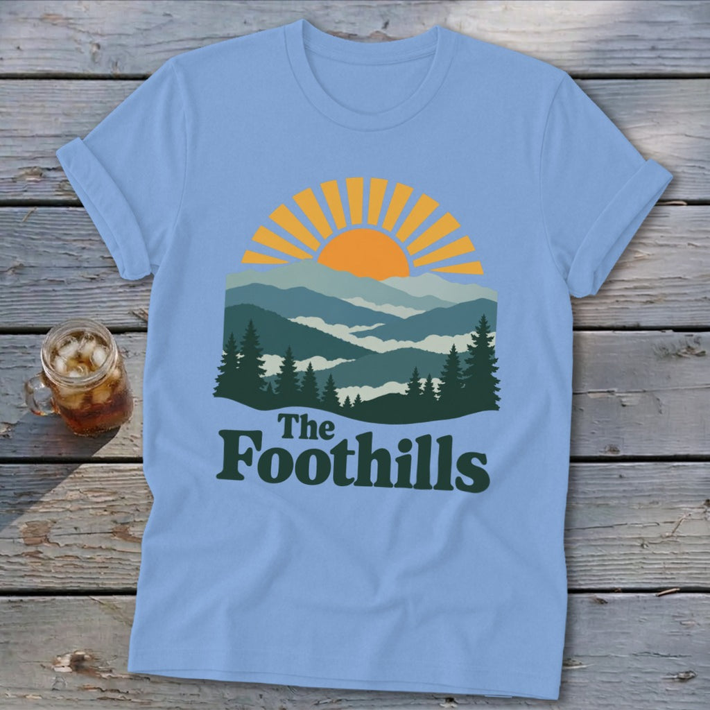 Light blue t-shirt with mountain and sun graphic and "The Foothills" text.