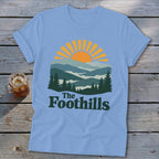 Light blue t-shirt with mountain and sun graphic and "The Foothills" text.