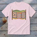 Light pink t-shirt featuring a graphic of three patchwork quilts hanging on a clothesline outdoors.