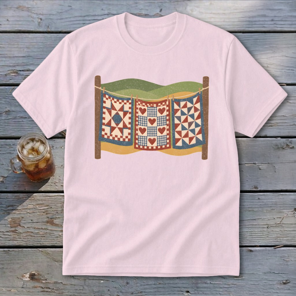 Light pink t-shirt featuring a graphic of three patchwork quilts hanging on a clothesline outdoors.