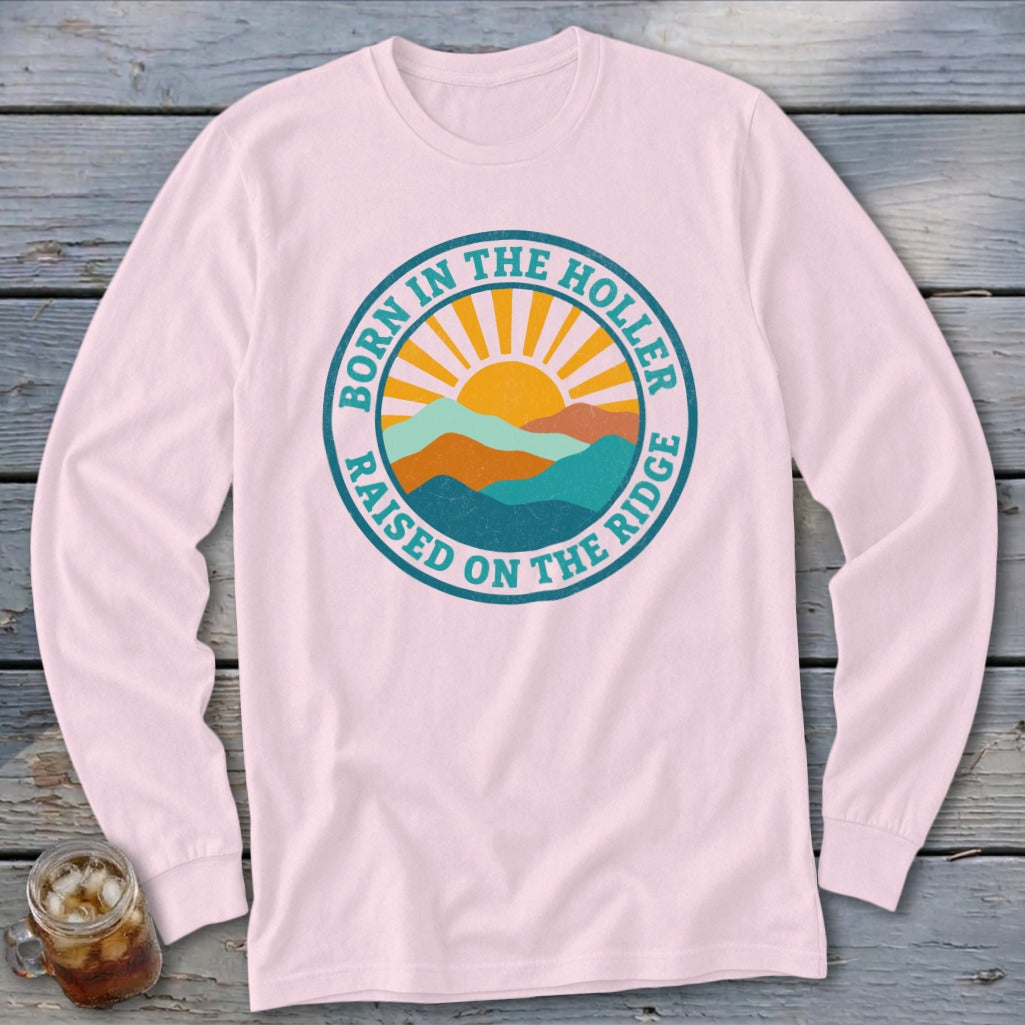 A light pink long-sleeve t-shirt features a circular retro mountain and sun design with the text Born in the Holler Raised on the Ridge.