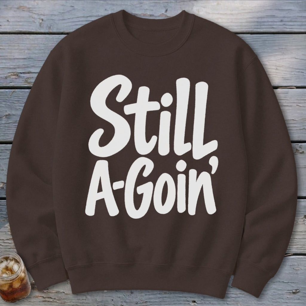 Brown crewneck sweatshirt printed with white text reading Still A-Goin’ laid flat against a rustic wooden background.