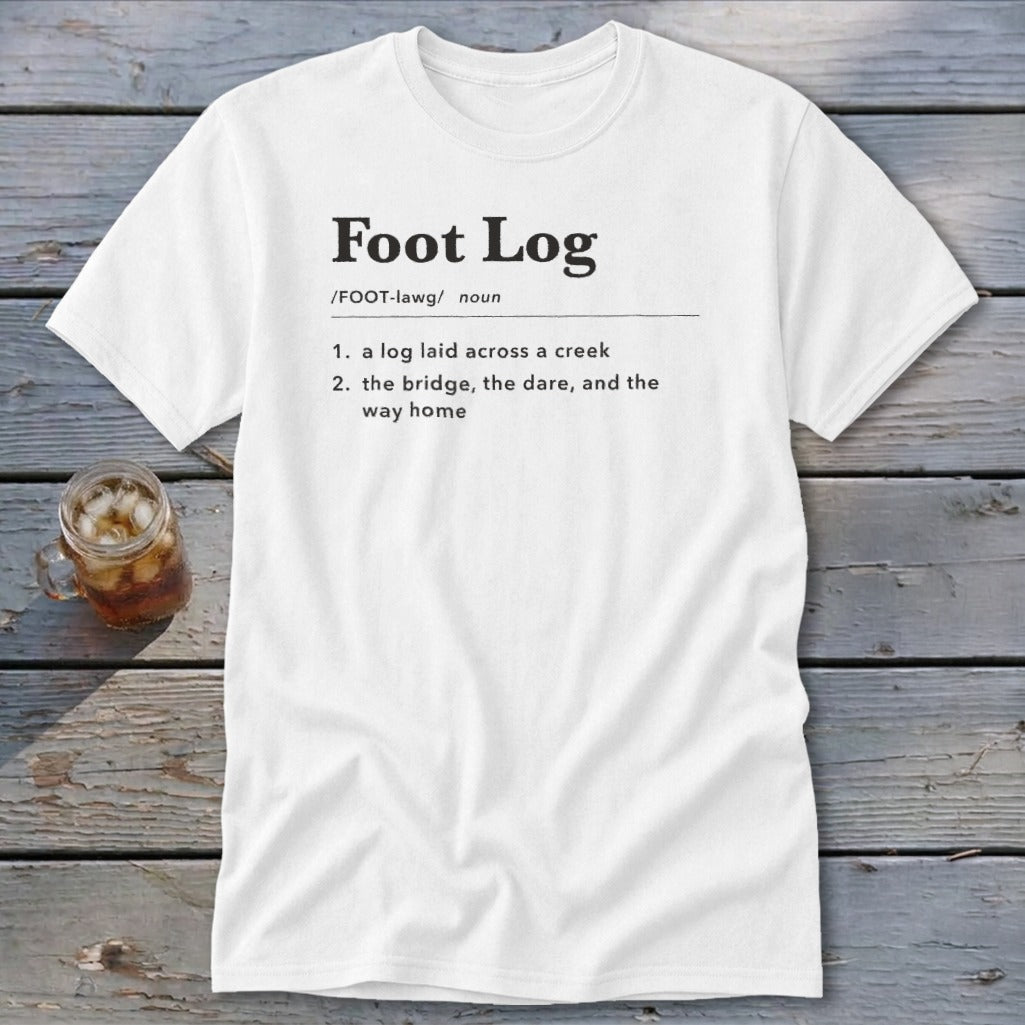 White t-shirt featuring dictionary-style text defining Foot Log laid across a wooden background.