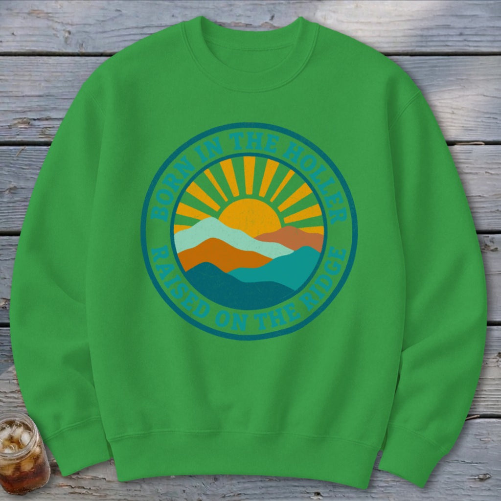 A green crewneck sweatshirt featuring a circular retro graphic with mountains, a sun, and text that reads BORN IN THE HOLLER RAISED ON THE RIDGE.
