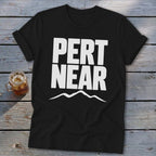 A black crewneck t-shirt featuring the white text PERT NEAR above a mountain range graphic, laid on a weathered wooden surface.